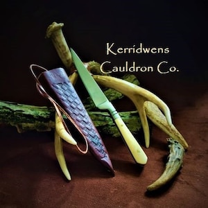 May include: A small, white-handled knife with a leather sheath and a wooden handle. The knife is resting on a piece of wood with two antlers. The text "Kerridwens Cauldron Co." is visible in the image.