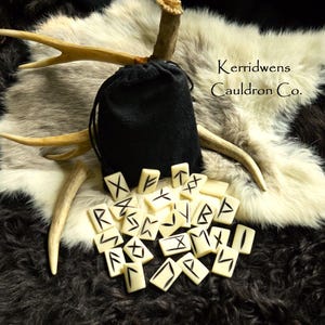 May include: A set of 25 white rune stones with black markings, arranged on a white fur surface. The runes are in a black velvet bag with a drawstring closure. The bag is sitting on a pair of antlers. The text "Kerridwens Cauldron Co." is visible in the upper right corner of the image.