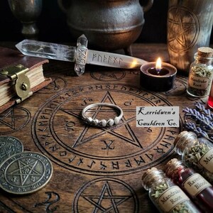 May include: A silver ring featuring a series of small moon phases along the band, resting on a wooden surface engraved with a pentagram and runic symbols. The ring is surrounded by a crystal dagger, a lit candle, and small glass vials of herbs.