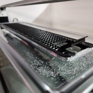 May include: A close-up of a black plastic grate over a clear glass aquarium tank. The grate is designed to prevent fish from jumping out of the tank. The water in the tank is clear and there are small bubbles visible.