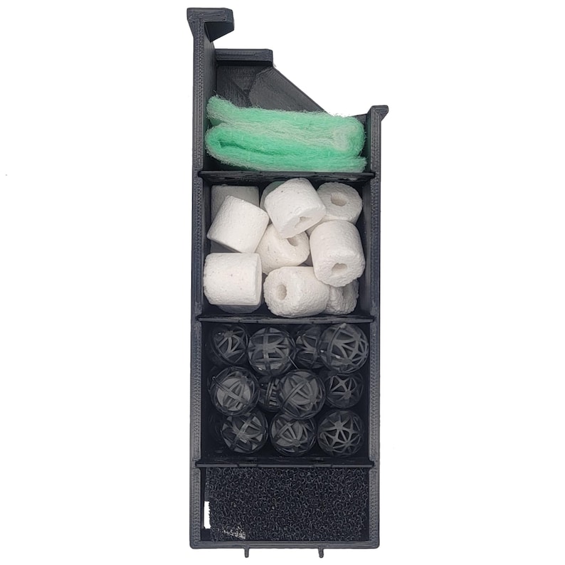 Op de afbeelding: Een zwarte plastic filtercartridge met vier compartimenten. Het bovenste compartiment bevat een groene filterpad, het tweede compartiment bevat witte keramische filtermedia, het derde compartiment bevat zwarte bioballen en het onderste compartiment bevat een zwarte sponsfilterpad.
