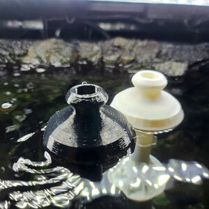 Puede incluir: Dos tapones de botella impresos en 3D, uno negro y otro blanco, flotando en el agua. El tap&oacute;n negro tiene una parte superior lisa y redondeada y una base texturizada y acanalada. El tap&oacute;n blanco tiene una parte superior lisa y redondeada y una base lisa y redondeada.