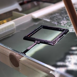 May include: A black plastic frame with a clear plastic screen, floating in a clear liquid. The frame is attached to a black handle.