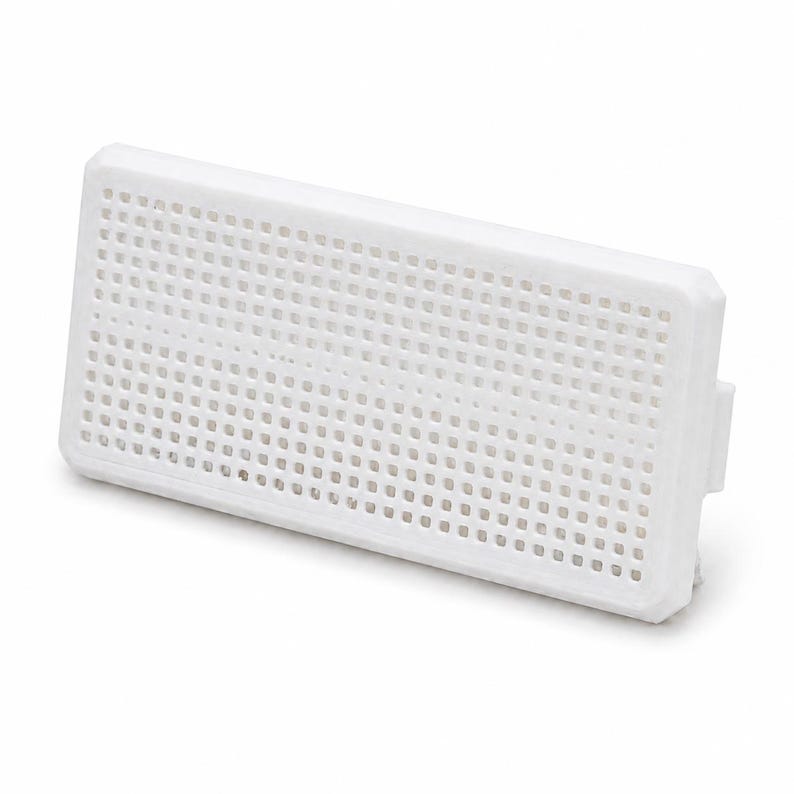 May include: A rectangular white plastic component featuring a uniform grid of small square perforations across its front surface. The object has a solid frame and a protruding tab on the right side, designed for insertion into a larger assembly.