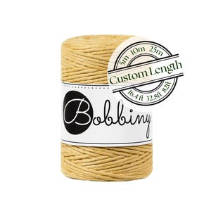 May include: A spool of mustard yellow cord wrapped in a white paper label featuring the text Bobbiny and a circular graphic with the words Custom Length and measurements 5m, 10m, 25m, 16.4ft, 32.8ft, and 82ft printed in black ink on the label.