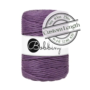 May include: A tightly wound spool of purple macrame cord. A white label wraps around the cord with the word "Bobbiny" in a script font. A circular sticker indicates custom lengths of 5m, 10m, and 25m, and 16.4 ft and 82 ft.