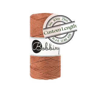 May include: A spool of terracotta-colored cord wrapped with a white paper label featuring the text Bobbiny and a circular graphic indicating Custom Length with options for 5m, 10m, 25m, 16.4ft, 32.8ft, and 82ft for various crafting projects.