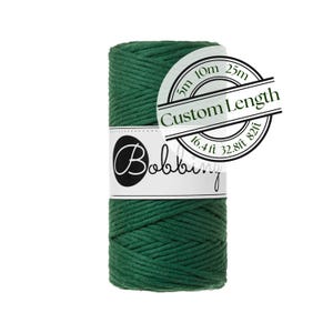 May include: A spool of dark green cord wrapped in a white paper label with the text Bobbiny and a circular graphic showing Custom Length with options for 5m, 10m, 25m, 16.4ft, 32.8ft, and 82ft. The cord has a braided texture and a solid color.