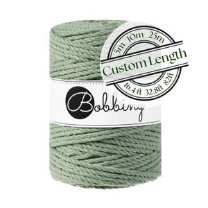 May include: A spool of sage green macrame cord wrapped in a white paper band with the brand name Bobbiny printed in black. A circular overlay displays the text Custom Length with measurements of 5m, 10m, 25m, 16.4ft, 32.8ft, and 82ft.