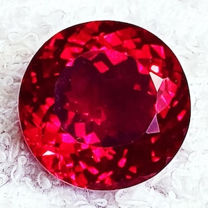Puede incluir: Una piedra preciosa redonda y facetada de un color rojo intenso y vibrante. La piedra presenta una estructura interna compleja con numerosas facetas geométricas reflectantes que crean un aspecto brillante y multidimensional.