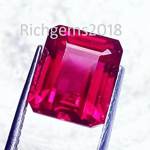 May include: A vibrant, emerald-cut red gemstone held by silver-toned metal tweezers. The stone features a faceted surface with deep crimson hues and high clarity. The text Richgems2018 is visible in the upper portion of the image background.