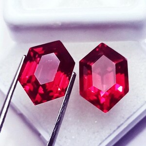 Puede incluir: Dos piedras preciosas rojas vibrantes con facetas hexagonales y corte brillante. Una piedra es sostenida por pinzas de metal plateado, mientras que la otra descansa cerca. Las piedras exhiben tonos carmesí profundos y bordes geométricos definidos.