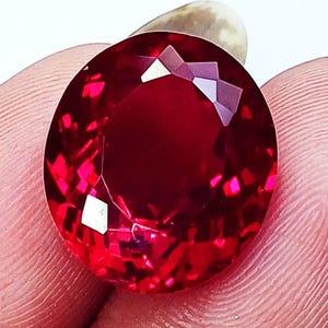 May include: A vibrant, oval-cut gemstone with a deep red colour and a faceted surface that reflects light. The stone displays high clarity and intense saturation, showcasing a brilliant, polished finish throughout its entire structure.