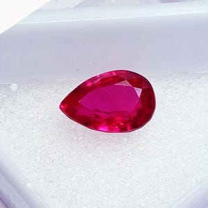 May include: A vibrant, deep pinkish-red gemstone cut into a pear shape with multiple facets that reflect light. The gemstone is displayed resting on a white, textured foam insert inside a small, white plastic storage container.