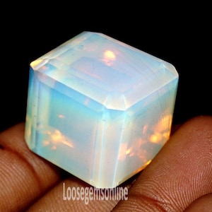 Puede incluir: Una gema de ópalo facetada en forma de cubo. La gema muestra un color base blanco lechoso con destellos iridiscentes azules y naranjas. La piedra se sostiene en una mano sobre un fondo negro.