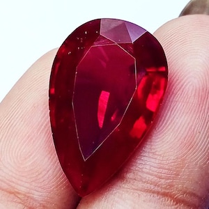 May include: A pear-shaped, faceted gemstone with a deep, vibrant red colour. The stone features a polished surface with multiple geometric facets that reflect light, creating a brilliant appearance. The gemstone is translucent and displays a rich crimson hue.