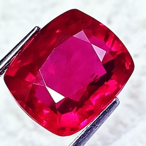 Peut inclure: Une pierre précieuse rouge taille coussin, maintenue par des pinces en métal argenté. La pierre présente une surface à facettes qui reflète la lumière, révélant une teinte cramoisie profonde et saturée avec une clarté interne nette.