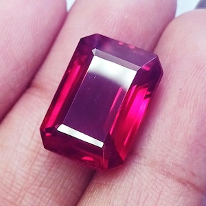 Puede incluir: Una gema de color rojo intenso y vibrante con un corte esmeralda rectangular. La piedra presenta facetas nítidas y limpias que crean una superficie altamente reflectante, mostrando un tono carmesí rico y saturado en toda su estructura.