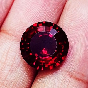 May include: A round, faceted gemstone with a deep red colour and a dark, opaque centre. The stone features a complex, geometric pattern of light-reflecting facets around its perimeter, creating a vibrant, sparkling effect against the dark core.
