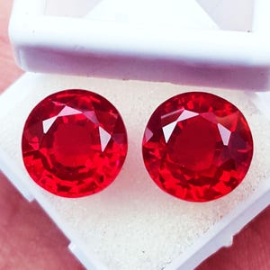 May include: Two round, faceted red gemstones displayed side by side within a white plastic storage box. The stones exhibit a vibrant, saturated crimson hue with clear geometric facets that reflect light across their circular surfaces.