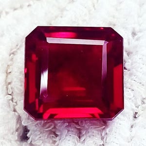 May include: A square-cut gemstone with a deep, vibrant red colour and faceted surfaces that reflect light. The stone features sharp, clean edges and a polished finish, displaying a rich, saturated crimson hue throughout its transparent structure.