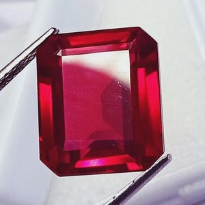 Op de afbeelding: Een levendige, dieprode edelsteen met een rechthoekige smaragdgeslepen vorm, vastgehouden door een metalen pincet. De steen heeft scherpe, heldere facetten die het licht reflecteren, wat zorgt voor een briljante, transparante uitstraling.