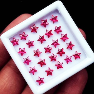 May include: A collection of small, star-shaped gemstones in a white tray. The gemstones are a vibrant shade of red, with multiple facets that catch the light. The tray is held against a dark background.