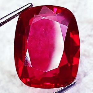 Op de afbeelding: Een levendig rode edelsteen met een kussenslijpsel, voorzien van meerdere facetten die het licht reflecteren. De steen wordt aan de boven- en onderkant vastgehouden door een metalen gereedschap, wat de dieprode kleur en textuur toont.