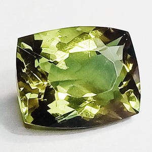 May include: A faceted, cushion-cut gemstone with a blend of green and brown hues. The gem's surface reflects light, showcasing its multiple facets and internal clarity. The stone is set against a white background.