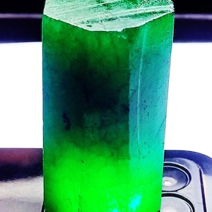 May include: A vibrant, emerald-green gemstone with a hexagonal shape. The gem has a polished top and a gradient of color, transitioning from a deep, dark green to a bright, glowing green at the base. The gemstone is displayed on a dark surface.