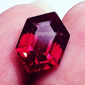 Puede incluir: Una gema facetada de color rojo intenso con forma geométrica. La gema tiene una superficie pulida que refleja la luz, creando reflejos y sombras. El texto "loosegemsonline" es visible en la gema. La gema está sujeta por una persona.