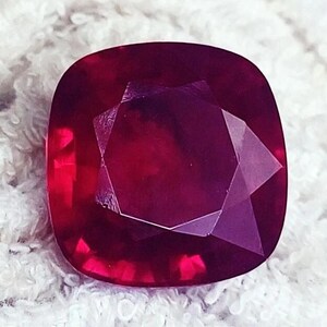 Puede incluir: Una piedra preciosa de color rojo intenso, con talla cojín y una superficie facetada que refleja la luz. La piedra presenta una forma cuadrada con esquinas redondeadas y un tono carmesí vibrante y saturado con facetas geométricas.