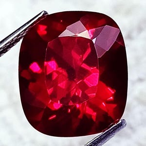 Puede incluir: Una piedra preciosa de corte cojín con un color rojo intenso y vibrante y una superficie facetada que refleja la luz. La piedra está sujeta por pinzas de metal en las esquinas superior izquierda e inferior derecha, mostrando su claridad.