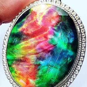 May include: An oval pendant featuring a vibrant, iridescent gemstone with swirling patterns of red, green, blue, and yellow. The stone is set in a polished silver-toned metal frame with a decorative, textured border and a top bail loop.