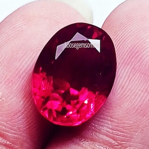 May include: An oval-cut, deep red gemstone with a faceted surface that reflects light. The stone features a white watermark overlay with the text "loosegemsonline" printed across the upper portion of the gem's surface.