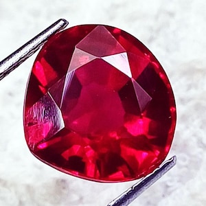 May include: A faceted, heart-shaped ruby gemstone. The ruby is a deep, vibrant red color and is held by a pair of silver tweezers. The gemstone has multiple facets that reflect light, creating a sparkling effect.