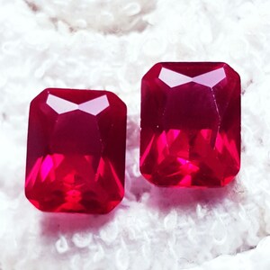 Puede incluir: Dos piedras preciosas de color rojo intenso y vibrante con un corte esmeralda rectangular. Cada piedra presenta múltiples facetas que reflejan la luz, creando una apariencia brillante y clara con bordes afilados y una superficie pulida.