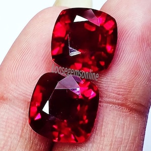 May include: Two cushion-cut, deep red gemstones with faceted surfaces that reflect light. The stones are positioned one above the other, and the text "Loosegemsonline" is printed in black across the center of the two stones.