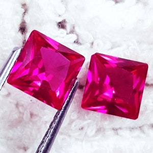Op de afbeelding: Twee levendige, vierkant geslepen fuchsia-roze edelstenen met gefacetteerde oppervlakken. De ene edelsteen wordt vastgehouden door een metalen pincet, terwijl de andere ernaast ligt en een briljante, reflecterende uitstraling heeft.