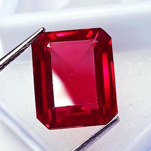 Puede incluir: Una gema roja de corte rectangular sostenida por unas pinzas metálicas. La piedra presenta una superficie facetada que refleja la luz, mostrando un tono carmesí profundo y saturado con bordes claros y un aspecto pulido y transparente.