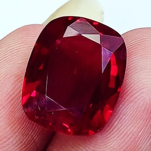 May include: A vibrant, deep red gemstone with a cushion cut, featuring multiple triangular and trapezoidal facets that reflect light. The stone displays a rich, saturated crimson hue throughout its transparent structure, showcasing precise geometric cuts.