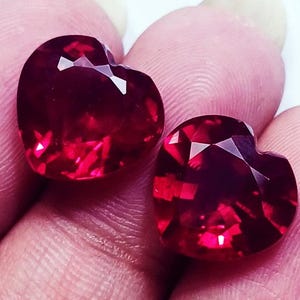 May include: Two heart-shaped, faceted, deep red gemstones. The gems have a brilliant, reflective surface, catching the light with multiple facets. The stones are held between fingers, showcasing their size and color.