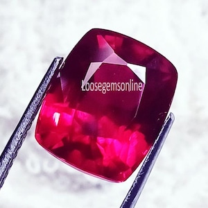 Peut inclure: Une pierre précieuse rouge foncé taille coussin avec une surface à facettes qui reflète la lumière. La pierre est tenue par des pinces en métal foncé et porte le texte Loosegemsonline imprimé en blanc sur le centre de la surface supérieure.