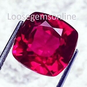 May include: A vibrant, cushion-cut red gemstone held by metal tweezers. The stone features a faceted surface that reflects light, creating deep crimson and bright pink highlights. The text Loosegemsonline is visible across the top of the stone.