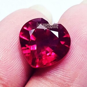 May include: A heart-shaped, faceted, deep red gemstone. The gem is held between fingers, showcasing its brilliant cut and vibrant color. The surface reflects light, highlighting the facets and the gem's clarity. The text "loosegemsonline" is visible on the gem.
