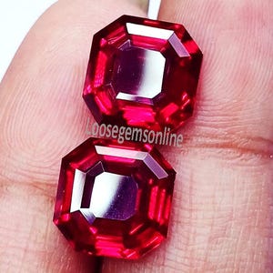 Könnte beinhalten: Zwei leuchtend rote, achteckig geschliffene Edelsteine mit facettierten Oberflächen, die das Licht reflektieren. Die Steine sind übereinander angeordnet und zeigen einen tiefen Karmesinton sowie scharfe, geometrische Kanten. Der Text "Loosegemsonline" ist zwischen ihnen zu sehen.