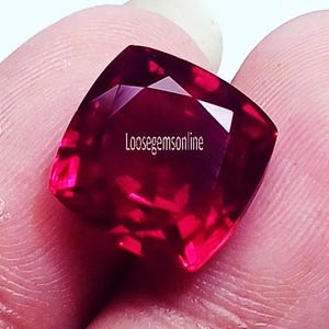 May include: A faceted, cushion-cut gemstone in shades of deep red and pink. The gem is held between fingers, with the text "loosegemsonline" visible on the surface. The gem's facets reflect light, creating a sparkling effect.