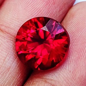 May include: A round, faceted gemstone with a vibrant, deep red colour. The stone features a complex pattern of geometric cuts that create internal light reflections and shadows, resulting in a brilliant, sparkling appearance across its surface.