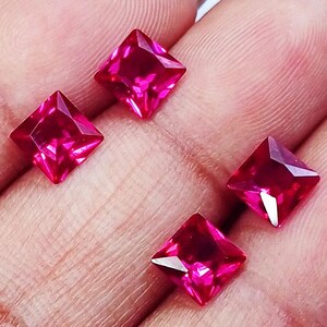 May include: Four square-cut, vibrant pinkish-red gemstones with faceted surfaces. Each stone features a pointed top and sharp edges, reflecting light to create a brilliant, translucent appearance. The stones are arranged in a loose cluster.