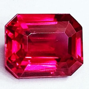 Peut inclure: Une pierre précieuse rectangulaire aux facettes d'un rouge profond et saturé. La pierre présente une surface taillée qui reflète la lumière, créant des reflets brillants et des ombres sombres sur sa structure polie, géométrique et transparente.